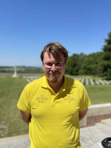 Roman poses in a yellow CWGF Guides polo shirt. CWGC headstones and cross of sacrifice are visible in the background.
