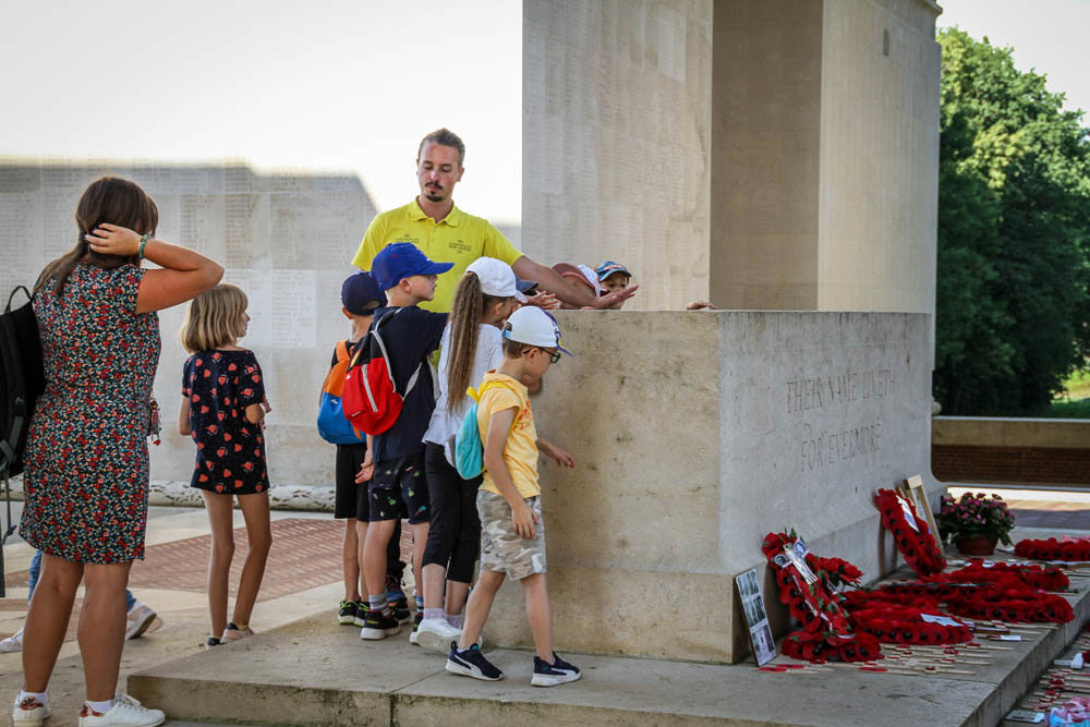 CWGF guide speaks to children and their parent on the Stone of Remembrance at Thiepval Memorial to the Missing. Lots of red poppy wreaths have been laid on the stone.